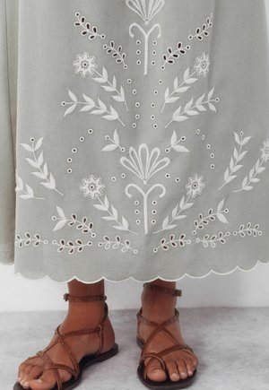 Jupe grise avec broderie florale blanche et ourlet festonné, portée avec des sandales plates marron à brides sur un sol clair.