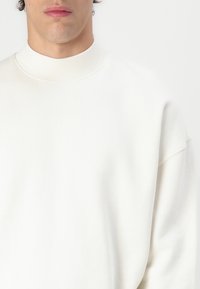Sweatshirt blanc avec un col rond haut et des épaules tombantes, fabriqué en tissu doux avec une texture lisse et des coutures minimales.