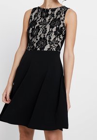 Robe noire avec un corsage en dentelle ajusté présentant des motifs floraux, sans manches et une jupe évasée au fini texturé.