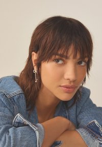 Jachetă din denim cu mânecile suflecate, având o nuanță albastră. Prim-plan al unei persoane cu păr închis la culoare și cercei din argint cu accente de perlă.
