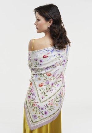 Tumšmataina sieviete ar ziedu raksta šalli pār pleciem un dzeltenu kleitu, skatās sāniski uz baltu fonu.