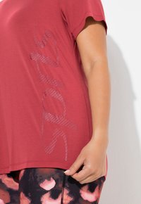 Czerwona koszulka z krótkim rękawem z teksturowanym nadrukiem „sports” w jaśniejszym odcieniu po boku, zestawiona z legginsami we wzory w odcieniach różu i czerni.