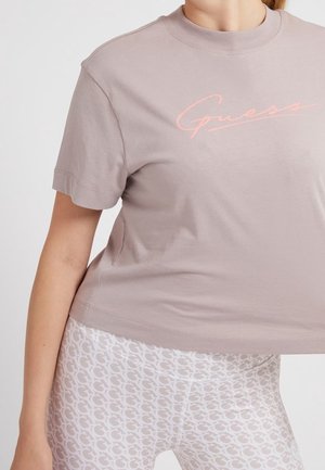 Vrouw die een licht mauve Guess T-shirt en witte leggings met een herhalend G-patroon draagt, poserend tegen een effen achtergrond.