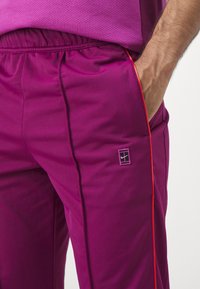 Pantaloni sportivi viola con vita elasticizzata, dotati di profilature arancioni sui lati e un piccolo logo sulla coscia sinistra. Texture liscia.