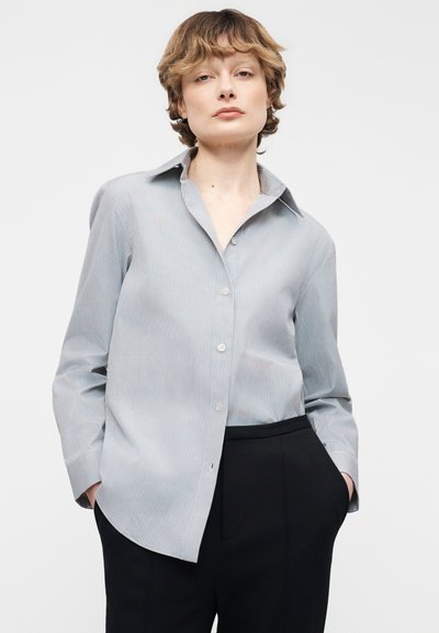 Νεαρό άτομο με κοντά μαλλιά που φοράει ανοιχτό μπλε πουκάμισο με ρίγες και μαύρο παντελόνι, στέκεται με τα χέρια στις τσέπες μπροστά σε λευκό φόντο.