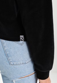 Schwarzes Samt-Sweatshirt mit gerippten Bündchen und Saum. Mit einem kleinen weißen Puma-Logo-Label auf der unteren linken Seite. Kombination mit hellblauen, ausgewaschenen Jeans.