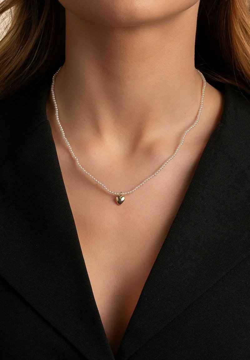 Kvinna som bär ett delikat pärlhalsband med en liten guldhjärtanhänge över ett svart V-ringat plagg.