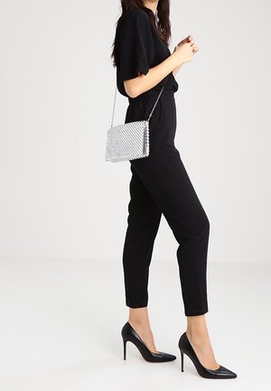 Černý overal s krátkými rukávy, zúženými nohavicemi a přiléhavým pasem; doplněný černými špičatými podpatky a stříbrnou kovovou kabelkou přes rameno.