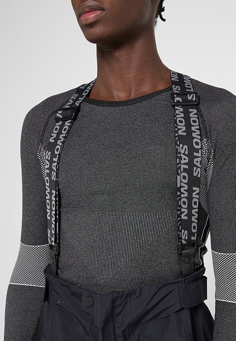 Zwart en grijs langmouwen top met een gestructureerd patroon. Voorzien van dikke zwarte verstelbare bandjes met de naam "Salomon" en zwarte broeken met bretels.