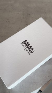 Caixa retangular branca feita de papel resistente, com o texto impresso em preto "MM6 Maison Margiela PARIS" na parte superior. Superfície lisa, design minimalista.