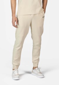 Pantaloni de trening bej cu croială conică, manșete elastice, buzunare laterale și un logo subtil. Asociați cu adidași albi cu accente bej deschis.