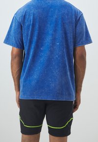 Modrá bavlněná trička s texturovaným povrchem, kombinovaná s černými šortkami s zeleným akcentovým šitím a těsným střihem.