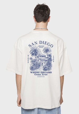Camiseta blanca de gran tamaño con estampado gráfico azul de palmeras y un edificio, con el texto "SAN DIEGO" y "DONDE LOS SUEÑOS Se Hacen Realidad".