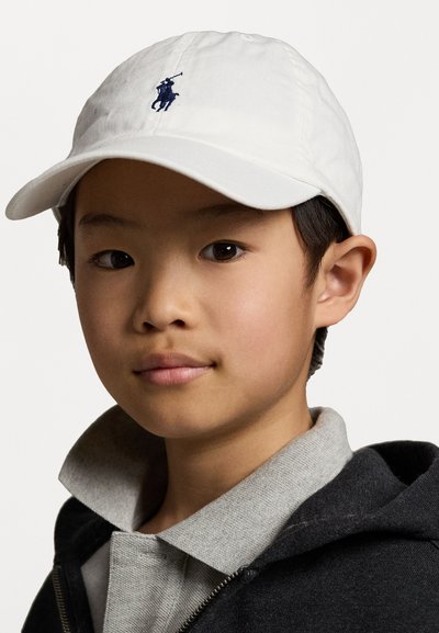 Witte baseballcap met een marineblauw geborduurd logo van een polo speler. Zacht materiaal, gebogen rand, iets scheef gedragen, gecombineerd met een grijze hoodie.