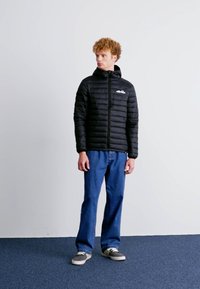 Zwarte geïsoleerde jas met een ritssluiting aan de voorkant, capuchon en geribde manchetten, gecombineerd met blauwe rechte jeans en grijze sneakers. Eenvoudig logo op de borst.