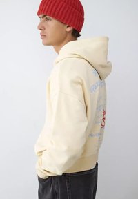 Sudadera con capucha color crema con cordón ajustable, bolsillos laterales y gráficos escritos a mano en azul y rojo. Combinada con un gorro de punto rojo acanalado, mostrando un look casual.