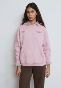 Sudadera con capucha rosa hecha de tela suave, con un bolsillo delantero y una camisa a rayas con cuello debajo. Detalles de texto en rojo.