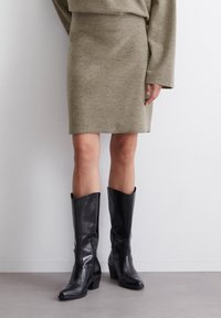 Jupe tricotée vert olive associée à des bottes en cuir noir arrivant jusqu'au genou avec un talon épais et un bout pointu. Texture lisse avec un minimum de détails.