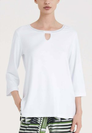 Donna che indossa una maglia bianca con maniche a 3/4 e scollo a goccia e pantaloni a righe neri, bianchi e verdi con fantasia.