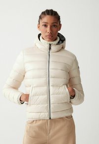 Giacca a vento leggera in crema, con texture trapuntata orizzontale, chiusura con zip e tasche. Design con cappuccio e silhouette aderente.