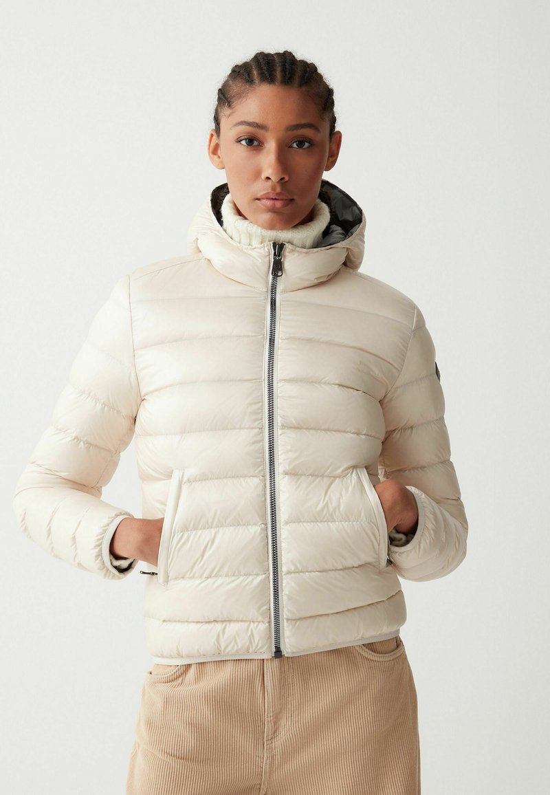 Giacca a vento leggera in crema, con texture trapuntata orizzontale, chiusura con zip e tasche. Design con cappuccio e silhouette aderente.