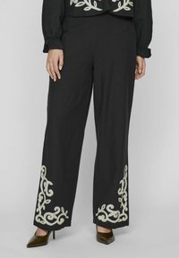 Pantalones negros de pierna ancha con intrincados bordados florales blancos en el dobladillo, confeccionados en una tela suave, complementados con tacones de punta.