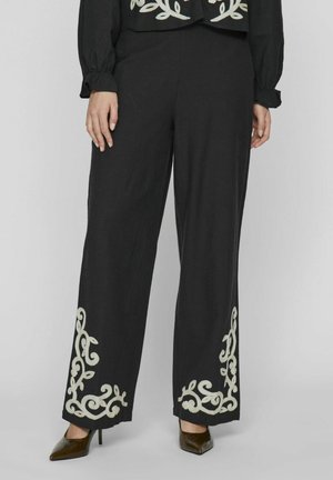 Pantalones negros de pierna ancha con intrincados bordados florales blancos en el dobladillo, confeccionados en una tela suave, complementados con tacones de punta.