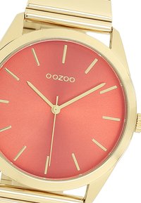 Montre-bracelet dorée avec un cadran rond en dégradé de corail, mettant en valeur des index et aiguilles dorés, ainsi qu'un bracelet plat en acier inoxydable.