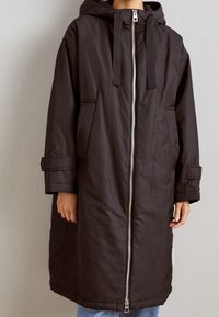 Manteau isolé noir avec capuche, fermeture éclair avant et poches. Présente une coupe longue et ample ainsi que des manches boutonnées pour un ajustement réglable.