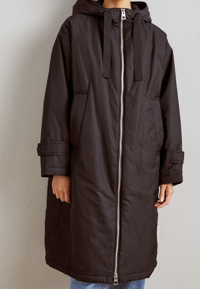 Manteau isolé noir avec capuche, fermeture éclair avant et poches. Présente une coupe longue et ample ainsi que des manches boutonnées pour un ajustement réglable.