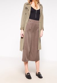 Olijfgroene lange cardigan over een zwart tanktopje, gecombineerd met wijd uitlopende bruine broeken. Zwarte loafers maken de outfit compleet en zorgen voor een strakke uitstraling.