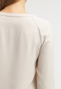 Haut de couleur crème avec une texture lisse, doté d'un large décolleté et de manches longues. Le dos présente des coutures nettes.