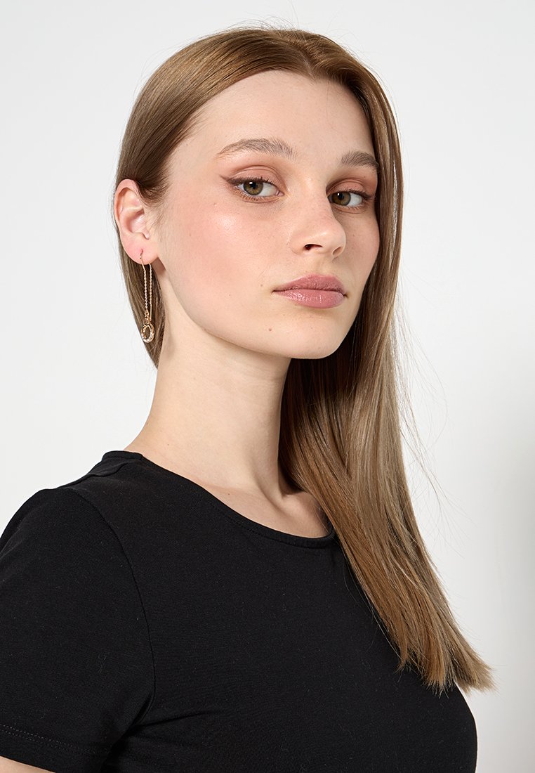 Boucles d'oreilles en or avec un accent circulaire, portées par un modèle avec des cheveux bruns raides et de longueur épaule, vêtu d'un t-shirt noir.