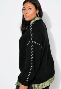 Schwarzer Strickpullover mit lockerer Passform, der weiße Nähte an den Ärmeln aufweist. Darüber trägt er ein grünes Tie-Dye-Shirt.