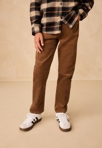 Pantalon en velours côtelé marron à coupe droite, assorti à une chemise en flanelle à carreaux noire et crème, et des baskets blanches avec des accents noirs.