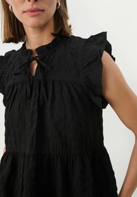 Schwarzes Kleid aus strukturiertem Stoff mit einem runden Hals, einer Bindung, rüschierten Ärmeln und einem leicht ausgestellten Rock.