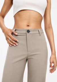Pantalon beige ajusté avec un tissu lisse, une fermeture à bouton à l'avant et des passants de ceinture. La coupe est droite et légèrement raccourcie à la taille.