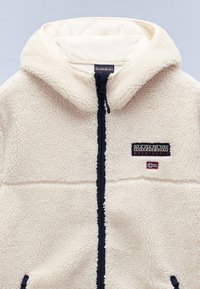 Veste polaire couleur crème avec capuche, fermeture éclair noire, patch logo Napapijri et petit drapeau norvégien sur la poitrine.