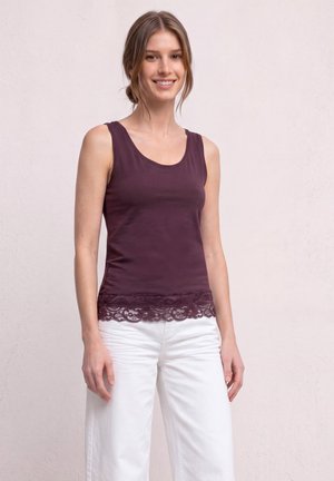 Jeune femme aux cheveux bruns attachés en chignon, portant un haut sans manches violet foncé avec un ourlet en dentelle et un pantalon blanc, debout devant un fond uni.