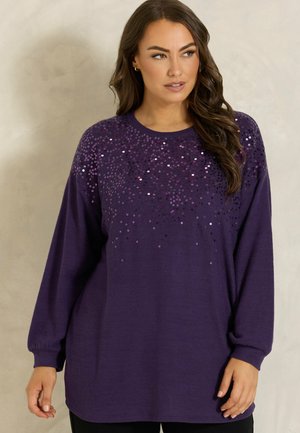 Femme portant un pull violet à manches longues avec des sequins brillants dispersés sur le haut devant et les manches, regardant légèrement sur le côté.