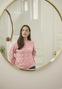 Rosa gestrickter Pullover mit Rautenmuster, Rundhalsausschnitt und langen Ärmeln. Im runden Spiegel betrachtet, mit einer Treppe und Pflanzen im Hintergrund.