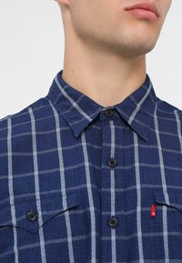 Chemise à carreaux bleue en tissu tissé, avec des lignes grises, un devant à boutons, deux poches poitrine et une étiquette rouge sur la poche gauche.