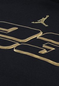 Hanorac din țesătură neagră cu litere metalizate aurii și logo-ul Jumpman, evidențiind o textură fină și elemente de design îndrăznețe.