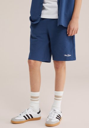 Osoba nosiace modré textúrované šortky s logom „Blue Ridge“, biele ponožky s béžovými pruhmi a biele tenisky Adidas s čiernymi pruhmi.