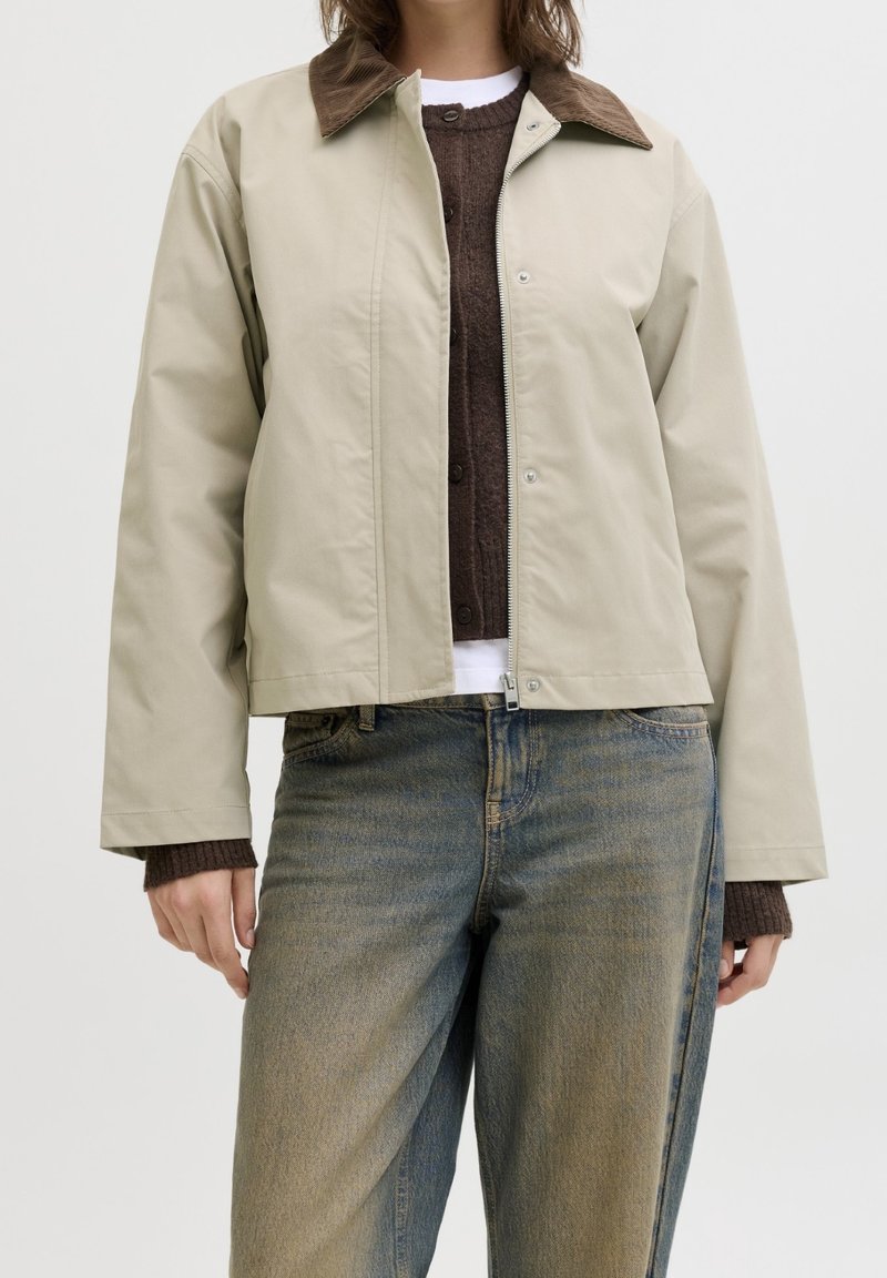 Personne portant une veste beige avec un col marron, un pull marron et un jean bleu délavé, visible du cou aux hanches sur un fond uni.