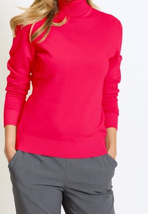 Femme portant un pull à col roulé rose vif et un pantalon gris, mains dans les poches, cadrée au niveau du cou et des cuisses.