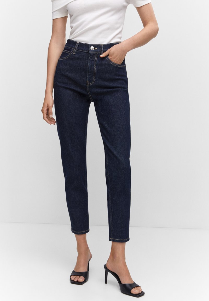 Femme portant un jean slim taille haute bleu foncé, un haut blanc à manches courtes et des mules à talons hauts ouvertes noires, se tenant debout avec une main dans la poche.