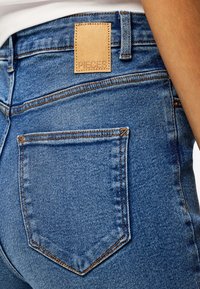 Gros plan sur la poche arrière d'un jean en denim bleu avec des coutures orange et une étiquette en cuir beige portant l'inscription « PIECES », porté par une personne en chemise blanche.