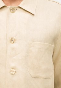 Μπεζ πουκάμισο με κουμπιά, τσέπη στο στήθος, γιακά και ορατά ράμματα σε ελαφρύ ύφασμα.