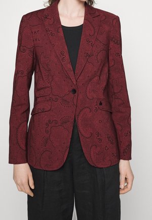 Vrouw draagt een nauwsluitend bordeauxrood colbert met zwarte paisley patronen over een zwart topje en losse zwarte broek, staand met ontspannen armen.